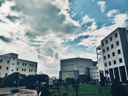 Several modern office buildings are positioned against a backdrop of a partly cloudy sky. The architectural structures are surrounded by greenery, including a well-maintained lawn and various plants, adding to the urban landscape.