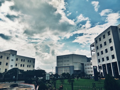 Several modern office buildings are positioned against a backdrop of a partly cloudy sky. The architectural structures are surrounded by greenery, including a well-maintained lawn and various plants, adding to the urban landscape.