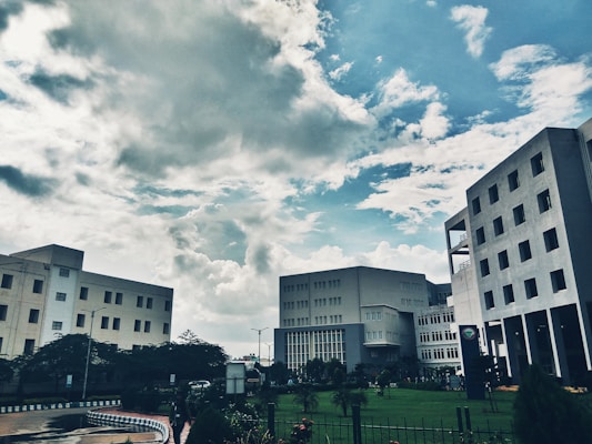 Several modern office buildings are positioned against a backdrop of a partly cloudy sky. The architectural structures are surrounded by greenery, including a well-maintained lawn and various plants, adding to the urban landscape.