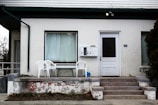 An exterior view of a white brick house featuring a small porch with two white plastic chairs and a table. The entryway is accessed by a set of concrete steps with visible peeling paint. The building has a large window with a curtain drawn across it and a white door with a black panel. A set of mailboxes labeled with numbers is mounted on the wall next to the door, and a house number '190' is visible. Sparse vegetation and a bare tree are seen around the porch area.