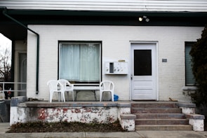 An exterior view of a white brick house featuring a small porch with two white plastic chairs and a table. The entryway is accessed by a set of concrete steps with visible peeling paint. The building has a large window with a curtain drawn across it and a white door with a black panel. A set of mailboxes labeled with numbers is mounted on the wall next to the door, and a house number '190' is visible. Sparse vegetation and a bare tree are seen around the porch area.