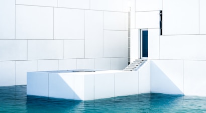 A minimalist architectural scene featuring a modern white building with geometric patterns. A staircase leads up to a door, and the structure seems to be partially surrounded by clear blue water.