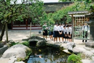 A group of students in uniforms stands by a small pond with koi fish, surrounded by greenery and stone features. They are in an outdoor setting with a traditional building and stone lantern nearby. The students appear to be engaged and observing the fish, creating an interactive and lively atmosphere.
