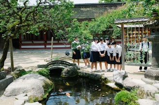 A group of students in uniforms stands by a small pond with koi fish, surrounded by greenery and stone features. They are in an outdoor setting with a traditional building and stone lantern nearby. The students appear to be engaged and observing the fish, creating an interactive and lively atmosphere.