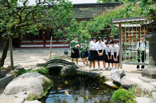 A group of students in uniforms stands by a small pond with koi fish, surrounded by greenery and stone features. They are in an outdoor setting with a traditional building and stone lantern nearby. The students appear to be engaged and observing the fish, creating an interactive and lively atmosphere.