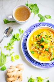 A vibrant bowl of soup sits on a blue and white patterned plate. The soup is filled with chunks of pumpkin, herbs, and leafy greens. Fresh ginger and coriander leaves are scattered around the plate on a light marble surface. A mug of golden broth and a spoon are placed nearby, adding to the culinary setting.