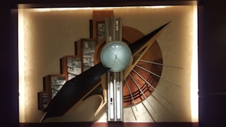 A large, abstract wall sculpture features geometric shapes, including a central sphere surrounded by a series of angular and curved elements. The composition includes a variety of metallic and earthy tones, with a prominent use of straight lines and radiating patterns. Architectural sketches are integrated into the background, adding depth and complexity to the piece.