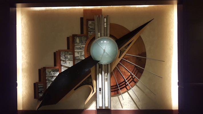 A large, abstract wall sculpture features geometric shapes, including a central sphere surrounded by a series of angular and curved elements. The composition includes a variety of metallic and earthy tones, with a prominent use of straight lines and radiating patterns. Architectural sketches are integrated into the background, adding depth and complexity to the piece.