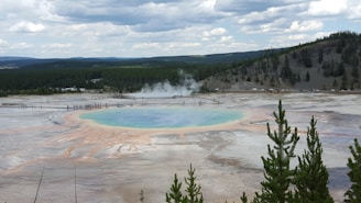 A large, vibrant geothermal spring is surrounded by a walkway with people observing. The spring features a blue and green center and orange and yellow rings due to mineral deposits and microbial life. In the background, there are pine forests on rolling hills under a partly cloudy sky.