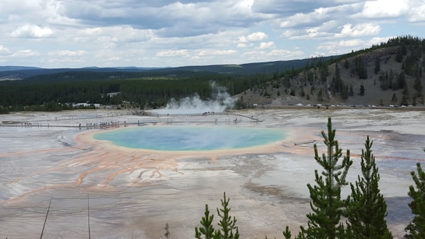 A large, vibrant geothermal spring is surrounded by a walkway with people observing. The spring features a blue and green center and orange and yellow rings due to mineral deposits and microbial life. In the background, there are pine forests on rolling hills under a partly cloudy sky.