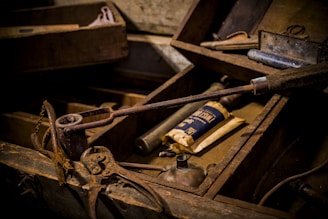 An assortment of old, rusted tools and items are displayed inside a wooden box with compartments. Among them, pliers, an oil can, a soldering iron, and a paper packet can be identified. The overall scene appears aged and worn, with a rustic, vintage ambiance.