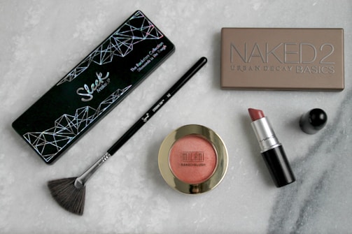 A collection of cosmetics is displayed on a marble surface, including an eyeshadow palette labeled 'Sleek Makeup,' a 'Naked 2' Urban Decay Basics palette, a Sigma brush, a Milani Baked Blush, and a MAC lipstick with its cap detached.
