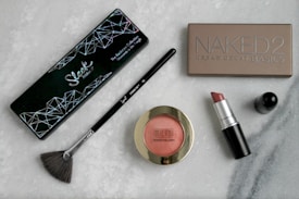 A collection of cosmetics is displayed on a marble surface, including an eyeshadow palette labeled 'Sleek Makeup,' a 'Naked 2' Urban Decay Basics palette, a Sigma brush, a Milani Baked Blush, and a MAC lipstick with its cap detached.