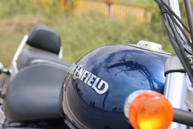 Close-up of a motorcycle fuel tank with the brand name visible, reflecting the sky and surrounding environment. The focus is on the shiny blue tank, while parts of the seat and handlebars are also visible.