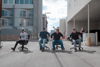 Four people are participating in a playful office chair race down an empty alley lined with buildings. Each person is seated on a swivel chair, propelling themselves forward using their feet. A woman stands in the background observing the race. The scene suggests a humorous and casual event.