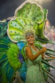 A person dressed in vibrant, elaborate carnival attire featuring bright green and blue hues. The costume includes a large headpiece adorned with jewels and intricate designs, and oversized decorative elements extend from the back, resembling feathers or leaves.