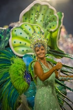 A person dressed in vibrant, elaborate carnival attire featuring bright green and blue hues. The costume includes a large headpiece adorned with jewels and intricate designs, and oversized decorative elements extend from the back, resembling feathers or leaves.