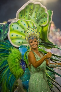 A person dressed in vibrant, elaborate carnival attire featuring bright green and blue hues. The costume includes a large headpiece adorned with jewels and intricate designs, and oversized decorative elements extend from the back, resembling feathers or leaves.