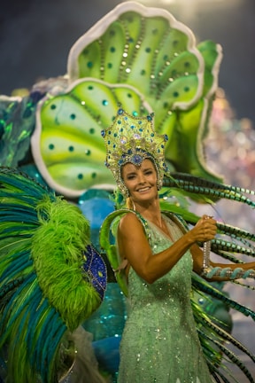 A person dressed in vibrant, elaborate carnival attire featuring bright green and blue hues. The costume includes a large headpiece adorned with jewels and intricate designs, and oversized decorative elements extend from the back, resembling feathers or leaves.
