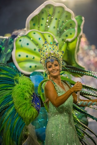 A person dressed in vibrant, elaborate carnival attire featuring bright green and blue hues. The costume includes a large headpiece adorned with jewels and intricate designs, and oversized decorative elements extend from the back, resembling feathers or leaves.