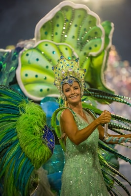 A person dressed in vibrant, elaborate carnival attire featuring bright green and blue hues. The costume includes a large headpiece adorned with jewels and intricate designs, and oversized decorative elements extend from the back, resembling feathers or leaves.