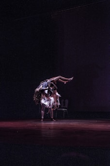A dancer in a well-lit stage scene performs an intricate lift, swinging another dancer over their shoulder. Both dancers wear costumes with flowing, fringe-like elements that add movement to the scene. The background is dark, focusing attention on the performers and the chair placed in the corner.
