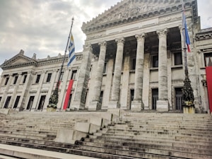 A grand neoclassical building with tall columns and intricate stone carvings dominates the scene. Two flags are prominently displayed, one of which is the flag of Uruguay. Wide stone steps lead up to the entrance, and the sky is overcast, casting a muted light over the structure.