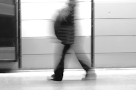 Blurred image of a person walking briskly past a light-colored, panel-lined wall. The motion creates a ghostly effect, with the person&rsquo;s form appearing stretched and translucent.