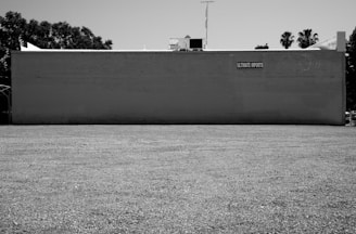 A large, flat building exterior painted in a light color with a simple sign reading 'Ultimate Imports' affixed near the top. The building has a plain, expansive gravel lot in front of it, surrounded by some trees in the background. The image is in black and white, suggesting a stark, utilitarian setting.