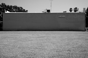 A large, flat building exterior painted in a light color with a simple sign reading 'Ultimate Imports' affixed near the top. The building has a plain, expansive gravel lot in front of it, surrounded by some trees in the background. The image is in black and white, suggesting a stark, utilitarian setting.