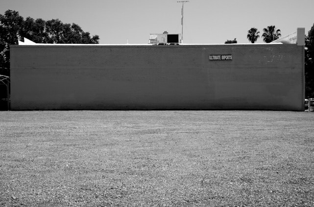A large, flat building exterior painted in a light color with a simple sign reading 'Ultimate Imports' affixed near the top. The building has a plain, expansive gravel lot in front of it, surrounded by some trees in the background. The image is in black and white, suggesting a stark, utilitarian setting.