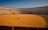 A person in a wheelchair is positioned at the center of an empty bullfighting ring. The ring is surrounded by red barriers and multi-tiered seating, with a clear blue sky in the background.