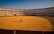 A person in a wheelchair is positioned at the center of an empty bullfighting ring. The ring is surrounded by red barriers and multi-tiered seating, with a clear blue sky in the background.