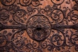 Intricate ironwork adorns a wooden surface, featuring ornate, swirling patterns with floral motifs. The craftsmanship showcases numerous spirals and leaf designs, centered around a circular iron ornament.