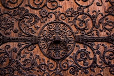 Intricate ironwork adorns a wooden surface, featuring ornate, swirling patterns with floral motifs. The craftsmanship showcases numerous spirals and leaf designs, centered around a circular iron ornament.