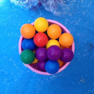A collection of colorful plastic balls is placed inside a pink container floating in a pool. The water is a vivid blue and the surface shows some ripples and splashes.
