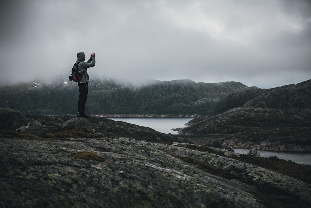 man standing on stones taking photo of mountain near body of water during foggy day