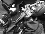 A pile of hockey equipment is spread out on the floor, including a helmet with a cage, gloves, elbow pads, and other protective gear. The items are arranged haphazardly, suggesting they might be ready for packing or just returned from use. The floor has a striped, textured surface, and the equipment is predominantly branded with names like Bauer and Alpha.