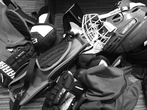A pile of hockey equipment is spread out on the floor, including a helmet with a cage, gloves, elbow pads, and other protective gear. The items are arranged haphazardly, suggesting they might be ready for packing or just returned from use. The floor has a striped, textured surface, and the equipment is predominantly branded with names like Bauer and Alpha.