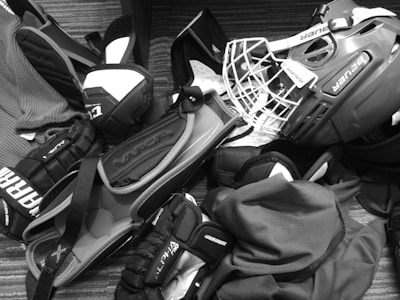 A pile of hockey equipment is spread out on the floor, including a helmet with a cage, gloves, elbow pads, and other protective gear. The items are arranged haphazardly, suggesting they might be ready for packing or just returned from use. The floor has a striped, textured surface, and the equipment is predominantly branded with names like Bauer and Alpha.