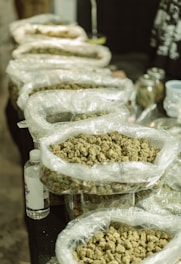 Several large plastic bags filled with what appears to be buds, possibly of a plant or herb, are arranged on a table. Next to the bags, there is a transparent bottle with a label, and in the background, glass jars and other containers can be seen.