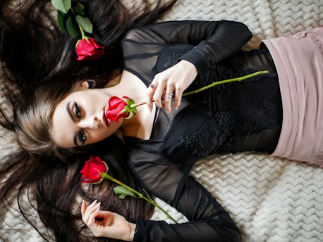 A woman lying on a textured blanket, surrounded by red roses. She wears a black lace top and holds a rose close to her face, with long dark hair spread around her.