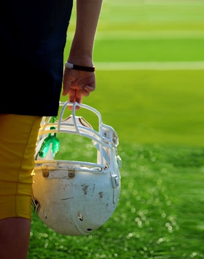 A person is holding a white football helmet by their side on a field. The individual is dressed in yellow and black athletic apparel. The grass is vivid green, with the sunlight creating a bright and fresh atmosphere.