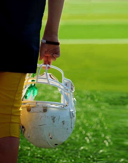 A person is holding a white football helmet by their side on a field. The individual is dressed in yellow and black athletic apparel. The grass is vivid green, with the sunlight creating a bright and fresh atmosphere.