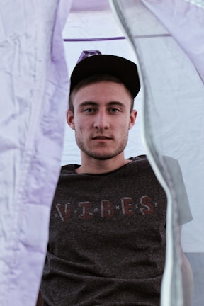 A person wearing a dark t-shirt with the word 'VIBES' is sitting inside a tent. The tent fabric is a light lavender color, creating a soft, diffused lighting. The person is wearing a dark cap and looking directly at the camera with a neutral expression.