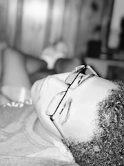 A person wearing glasses is reclining, seemingly relaxed and resting. The black and white image has a soft focus, and the lighting creates a serene atmosphere. The background is blurred, emphasizing the calmness of the moment.
