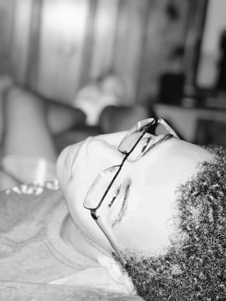 A person wearing glasses is reclining, seemingly relaxed and resting. The black and white image has a soft focus, and the lighting creates a serene atmosphere. The background is blurred, emphasizing the calmness of the moment.
