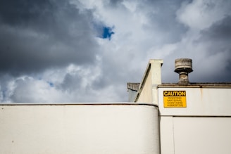 An industrial-style rooftop with a ventilation pipe against a backdrop of a cloudy sky. A prominent yellow caution sign warns of asbestos roofing material.