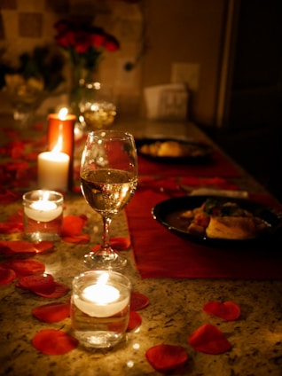 A cozy romantic dinner setup featuring candles, rose petals, and a custom gift box on a small table