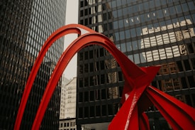 A vibrant red metal sculpture stands prominently against a backdrop of tall, reflective office buildings. The sculpture features sleek, curved lines and dynamic angles that create a striking contrast with the straight, grid-like patterns of the windows in the surrounding skyscrapers.